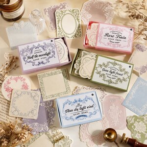 以下が含まれることがあります： Half the moonlight、Rose Train、Green light forest、Clear sky light windとラベルが貼られた長方形の箱に入った装飾用ステーショナリーセットのコレクション。様々な形の紙に、華やかな花の縁取りやヴィンテージ風のフレームが施されています。