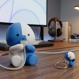 Può includere: Un supporto per cavo di ricarica a forma di personaggio blu e bianco con un portachiavi abbinato. Il supporto tiene un cavo di ricarica. Il portachiavi è una versione in miniatura del supporto. Su una scrivania di legno.