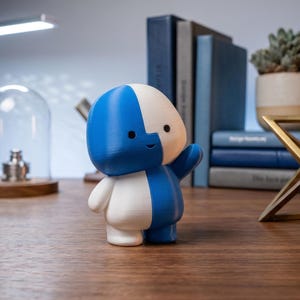 May include: A blue and white figurine of a cartoon character with a raised arm. The figure is standing on a wooden surface, with books and a succulent plant in the background. A decorative brass object is also visible.