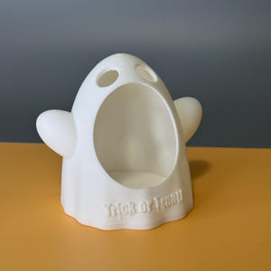 Puede incluir: Un recipiente blanco con forma de fantasma con una gran abertura y dos agujeros para los ojos más pequeños. El recipiente tiene pequeñas protuberancias en forma de brazo a cada lado. Las palabras "Trick or Treat" están grabadas en el borde inferior. El recipiente está sobre una superficie amarilla.