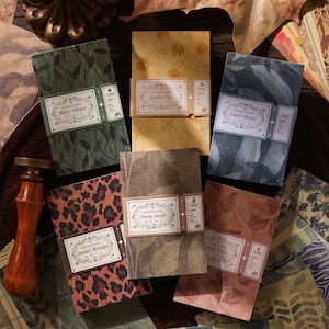 May include: Six rectangular packs of decorative scrapbook paper, each secured with a paper band featuring ornate labels. Designs include green leaf patterns, yellow florals, blue foliage, leopard print, brown botanical motifs, and pinkish leaf prints.