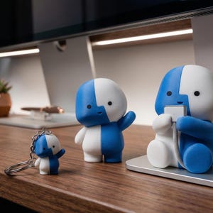 Puede incluir: Colección de figuras azules y blancas de varios tamaños, incluyendo un llavero, sobre una superficie de madera. Las figuras tienen un diseño de color dividido y una estética minimalista. Una figura sostiene un cable de carga.