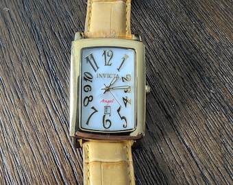 Puede incluir: Reloj Invicta Angel rectangular dorado con esfera crema y grandes números negros. El reloj tiene una correa de cuero amarilla con un patrón de cocodrilo. La esfera muestra la palabra "Invicta" y la palabra "Angel".