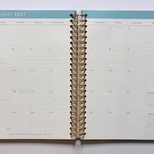 May include: An open spiral-bound planner showing a monthly calendar view for January 2027. The pages feature a grid layout with days of the week, a notes column, and small reference calendars for December 2026 and February 2027. The edges have colored tabs.