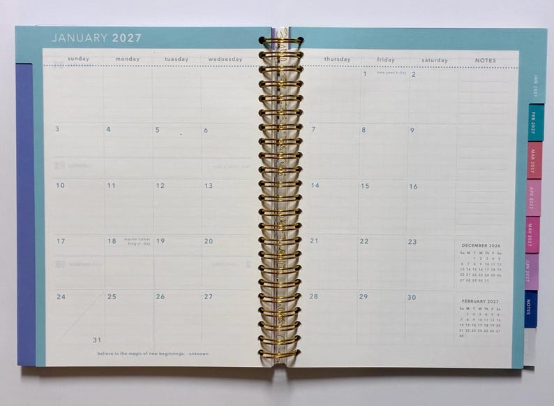 May include: An open spiral-bound planner showing a monthly calendar view for January 2027. The pages feature a grid layout with days of the week, a notes column, and small reference calendars for December 2026 and February 2027. The edges have colored tabs.