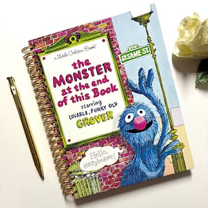 May include: A spiral-bound book titled The Monster at the End of This Book starring lovable, furry old Grover, featuring a blue monster character, a brick wall background, and a speech bubble saying Hello, everybodeee! next to a gold pen.