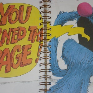 May include: A spiral-bound book page featuring a blue furry character with a pink nose and an open mouth. A yellow speech bubble on the left contains the orange text YOU TURNED THE PAGE! A yellow stream extends from the character's mouth toward the text.