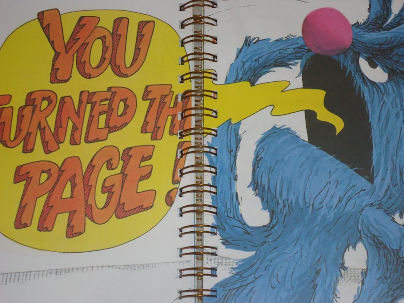 May include: A spiral-bound book page featuring a blue furry character with a pink nose and an open mouth. A yellow speech bubble on the left contains the orange text YOU TURNED THE PAGE! A yellow stream extends from the character's mouth toward the text.