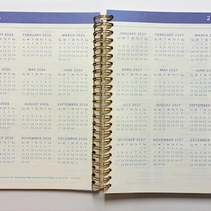May include: A spiral-bound planner open to a two-page spread displaying full-year calendars for 2026 and 2027. The pages feature cream-colored paper with blue text, gold-tone metal wire binding, and colorful index tabs on the right edge.