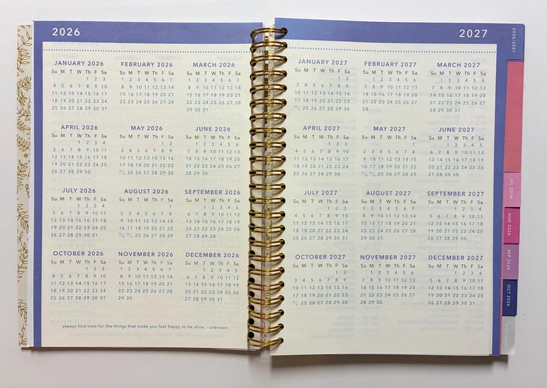May include: A spiral-bound planner open to a two-page spread displaying full-year calendars for 2026 and 2027. The pages feature cream-colored paper with blue text, gold-tone metal wire binding, and colorful index tabs on the right edge.