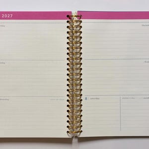 May include: An open spiral-bound planner showing the week of May 3, 2027, through May 9, 2027. The pages feature cream-colored paper with horizontal lines for writing, framed by bright pink borders. Gold-colored metal wire binding holds the pages.
