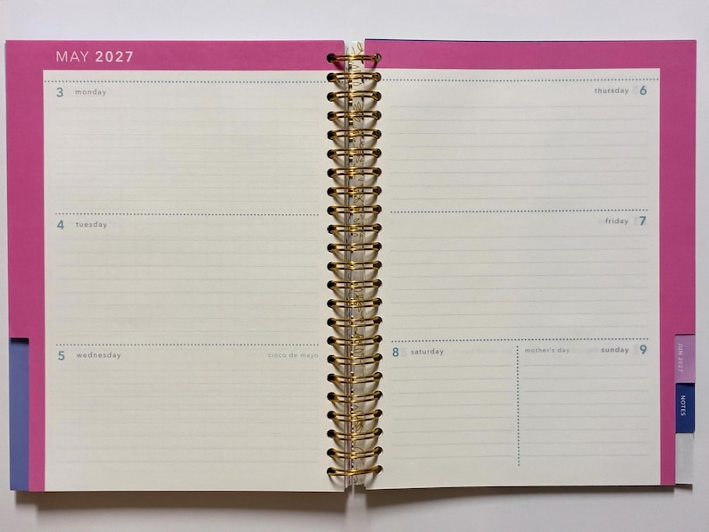 May include: An open spiral-bound planner showing the week of May 3, 2027, through May 9, 2027. The pages feature cream-colored paper with horizontal lines for writing, framed by bright pink borders. Gold-colored metal wire binding holds the pages.