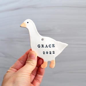 May include: A white ceramic goose ornament with a light orange beak and feet. The word "GRACE" and the year "2022" are printed in black on the goose's body. The ornament has a small hole at the top for hanging.