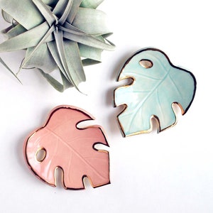 May include: Two ceramic monstera leaf dishes with gold trim. One dish is light blue and the other is pink.
