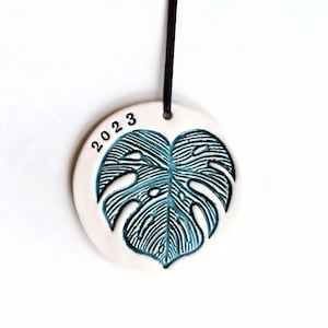 May include: White ceramic ornament with a teal green monstera leaf design and the year 2023 printed on the top. The ornament has a black string for hanging.