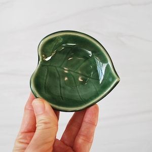 Puede incluir: Un peque&ntilde;o plato de cer&aacute;mica verde con forma de hoja. El plato tiene un acabado brillante y un dise&ntilde;o de hoja en relieve.