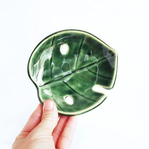 May include: A green ceramic dish shaped like a leaf with three holes. The dish is glazed and has a glossy finish.