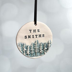 May include: A round, off-white ceramic ornament with the text "THE SMITHS" in black block letters. Below the text is a forest scene with blue-green trees. A black ribbon is attached for hanging.