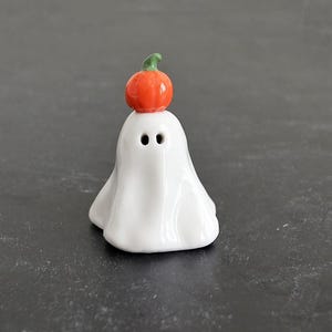 May include: A small white ceramic ghost figurine with a smooth, glossy finish. The ghost has two dark, circular eye holes and is topped with a miniature, vibrant orange pumpkin featuring a small green stem resting on its head.