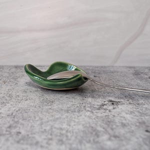 Puede incluir: Un reposacucharas de cer&aacute;mica verde con una cuchara en su interior. El reposacucharas tiene forma de hoja y un acabado brillante.