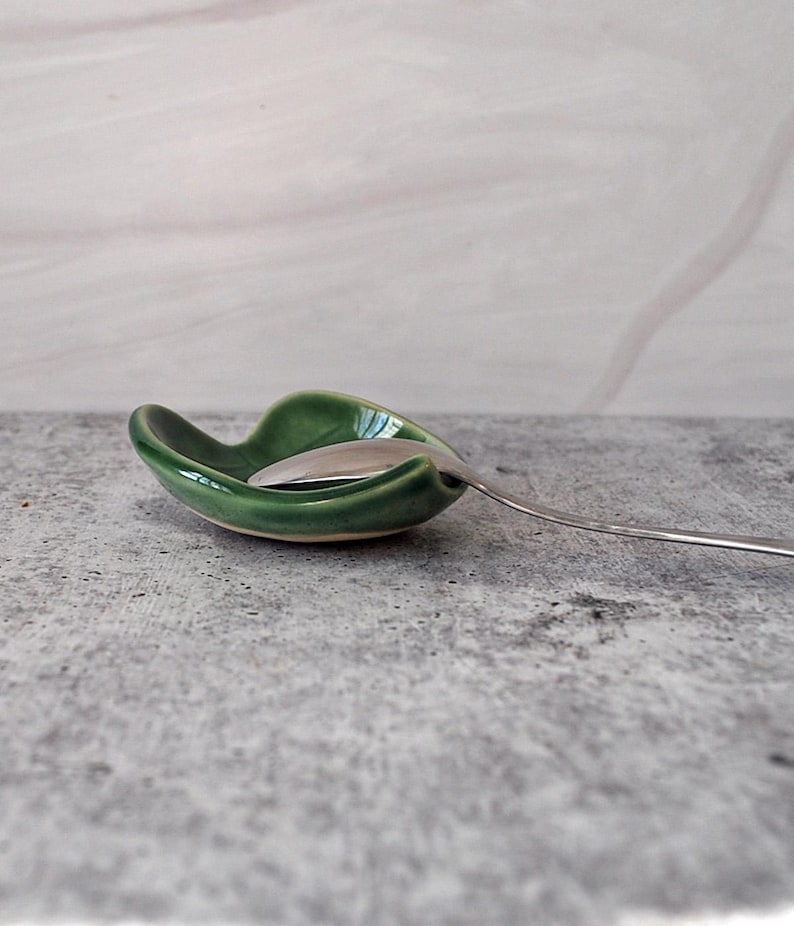 Puede incluir: Un reposacucharas de cer&aacute;mica verde con una cuchara en su interior. El reposacucharas tiene forma de hoja y un acabado brillante.