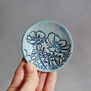 May include: A small, light blue ceramic dish with a raised design of two blue flowers. The dish is perfect for holding jewelry or other small items.