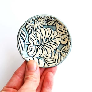 May include: A small, round ceramic dish with a blue glaze and a white embossed design of tropical leaves.