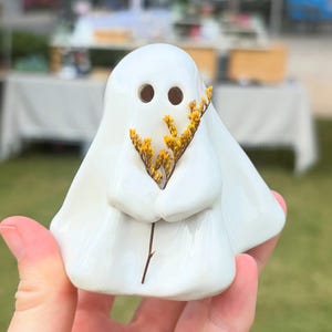May include: A white ceramic ghost figurine with a smooth, glossy finish, featuring two circular eye cutouts and holding a small, dried sprig of yellow flowers in its molded arms. The ghost has a rounded head and a draped, flowing body shape.