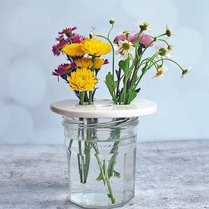 May include: A white ceramic flower frog with four holes sits atop a clear glass jar filled with water.  Yellow, pink, and white flowers are arranged in the frog.