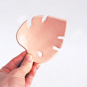 May include: A pink ceramic leaf-shaped dish. The dish has a hole in the center and a textured surface.