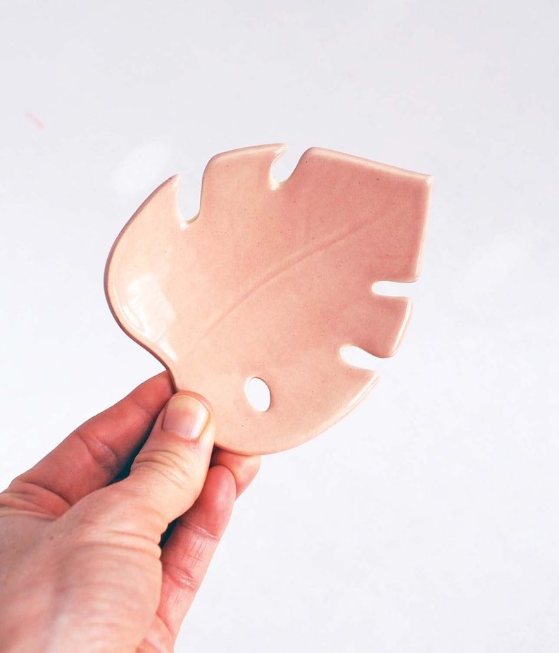 May include: A pink ceramic leaf-shaped dish. The dish has a hole in the center and a textured surface.