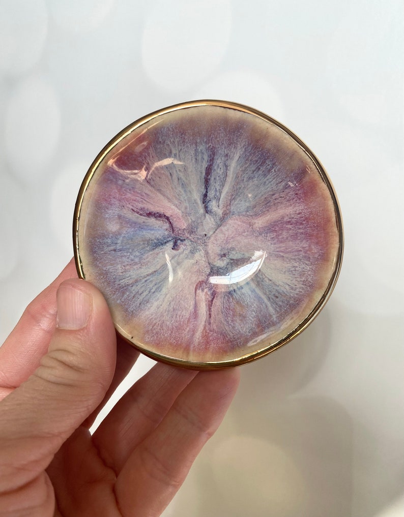 K&ouml;nnte beinhalten: Eine kleine, kreisf&ouml;rmige Schale mit einem wirbelnden, marmorierten Muster in sanften Violett-, Rosa- und Wei&szlig;t&ouml;nen. Die Innenfl&auml;che hat eine gl&auml;nzende, glatte Oberfl&auml;che, w&auml;hrend der &auml;u&szlig;ere Rand mit einer d&uuml;nnen, goldfarbenen Umrandung akzentuiert ist.