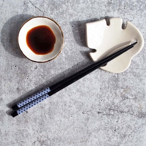 May include: A white ceramic leaf-shaped chopstick rest with a pair of black chopsticks with a blue and white geometric pattern. A small white bowl with a gold rim contains dark soy sauce.