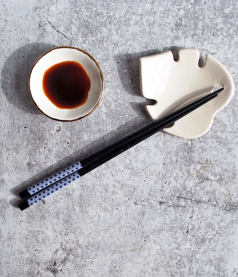 May include: A white ceramic leaf-shaped chopstick rest with a pair of black chopsticks with a blue and white geometric pattern. A small white bowl with a gold rim contains dark soy sauce.