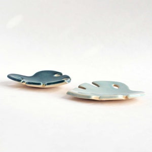 May include: Two ceramic leaf-shaped dishes, one in a light blue glaze and the other in a dark teal glaze. The dishes are made in the shape of a monstera leaf.