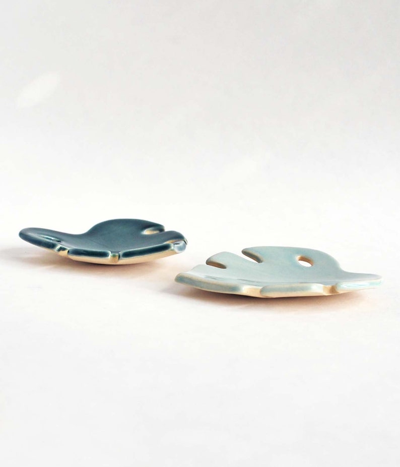 May include: Two ceramic leaf-shaped dishes, one in a light blue glaze and the other in a dark teal glaze. The dishes are made in the shape of a monstera leaf.