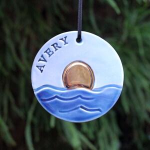 May include: A blue and white ceramic ornament with a gold sun and waves. The ornament is personalized with the name "AVERY".