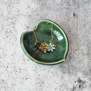 Puede incluir: Un collar de cadena dorada con un grupo de abalorios en forma de suculentas verdes, rojas y blancas. El collar se muestra en un plato de cer&aacute;mica verde en forma de hoja.