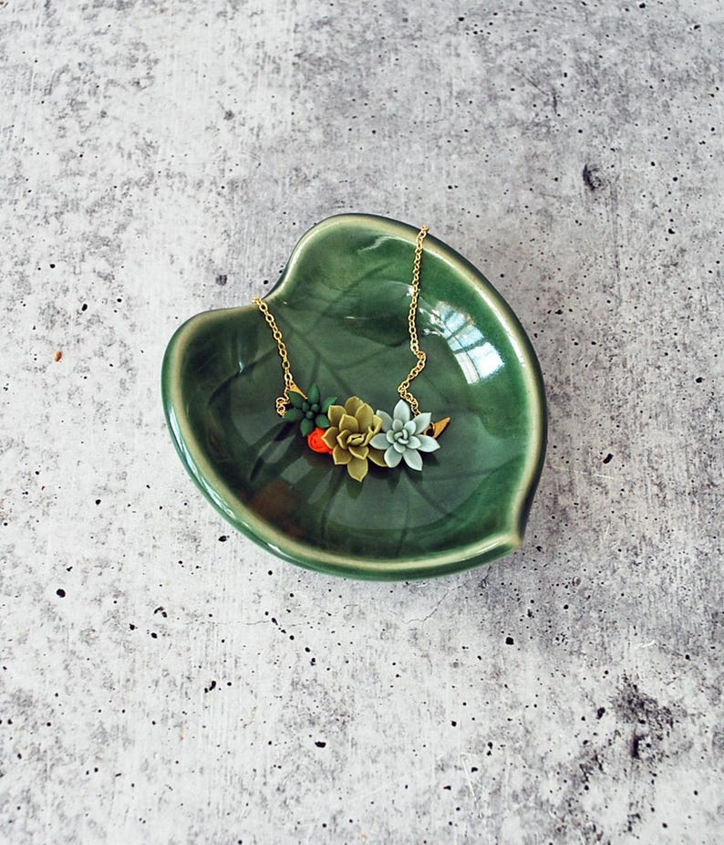 Puede incluir: Un collar de cadena dorada con un grupo de abalorios en forma de suculentas verdes, rojas y blancas. El collar se muestra en un plato de cer&aacute;mica verde en forma de hoja.