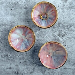 K&ouml;nnte beinhalten: Drei kleine, runde, flache Schalen mit einem wirbelnden, marmorierten Muster in Rosa-, Blau- und Wei&szlig;t&ouml;nen. Jede Schale hat einen glatten, goldfarbenen Rand. In einer der Schalen liegen zwei d&uuml;nne, goldfarbene Metallringe.