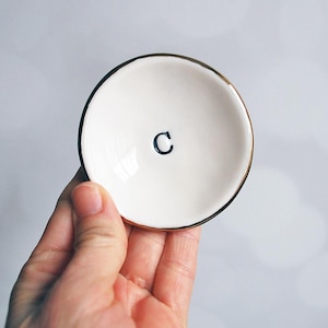 May include: A small, round, white ceramic trinket dish with a smooth, glossy finish. The center of the dish features a single, bold, black uppercase letter C. The outer rim of the dish is accented with a thin, metallic gold-colored edge.