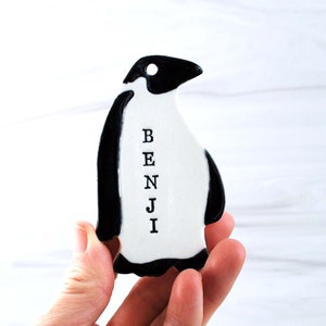 May include: A white ceramic penguin ornament with black accents. The penguin has the name "BENJI" written vertically on its chest.