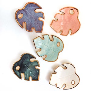 May include: Five ceramic monstera leaf dishes with gold rims. The dishes are in various colors, including purple, pink, blue, green, and white.