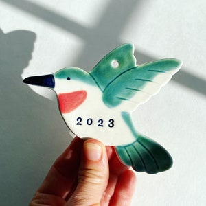 May include: A ceramic hummingbird ornament with a green and blue glaze. The hummingbird is in flight and has a red breast. The ornament is decorated with the year 2023.