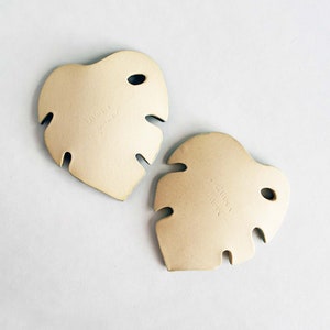 May include: Two beige ceramic monstera leaf shaped coasters. The coasters have a hole in the top and are marked with the words "Lauren" and "June".