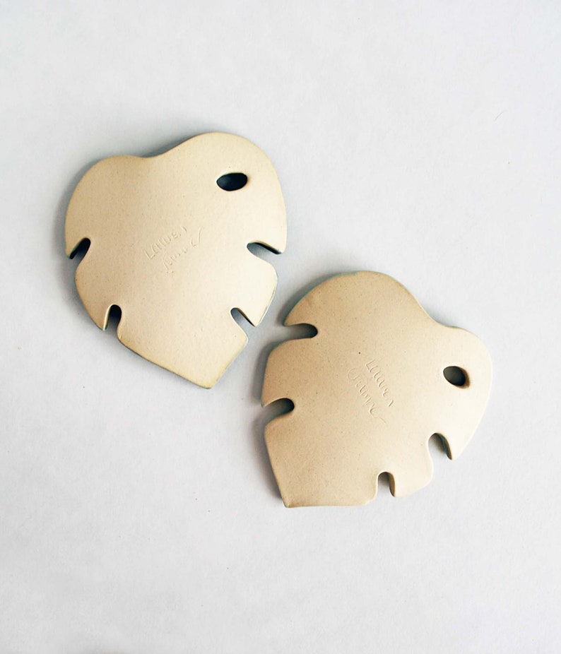 May include: Two beige ceramic monstera leaf shaped coasters. The coasters have a hole in the top and are marked with the words "Lauren" and "June".