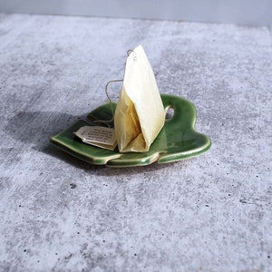 May include: A green ceramic leaf-shaped dish with a single tea bag resting on it. The tea bag is a pyramid shape and is made of white paper. The tea bag has a string attached to it and a small tag with the text "Your great happiness is based on the warmth of your heart".