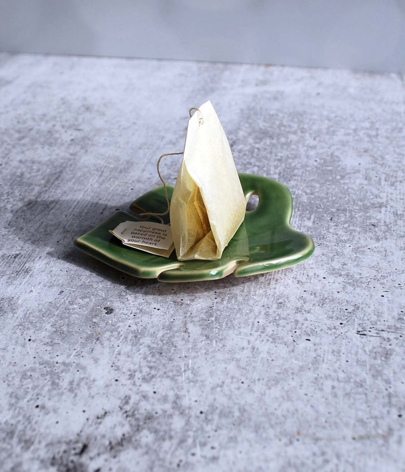 May include: A green ceramic leaf-shaped dish with a single tea bag resting on it. The tea bag is a pyramid shape and is made of white paper. The tea bag has a string attached to it and a small tag with the text "Your great happiness is based on the warmth of your heart".
