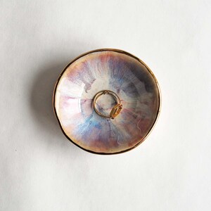 K&ouml;nnte beinhalten: Eine kleine, runde Schmuckschale aus Keramik mit einer schimmernden, irisierenden Glasur in zartem Rosa, Blau und Lavendel. Der Rand der Schale ist mit einem metallisch goldenen Akzent versehen. Ein goldfarbener Ring liegt in der Mitte.