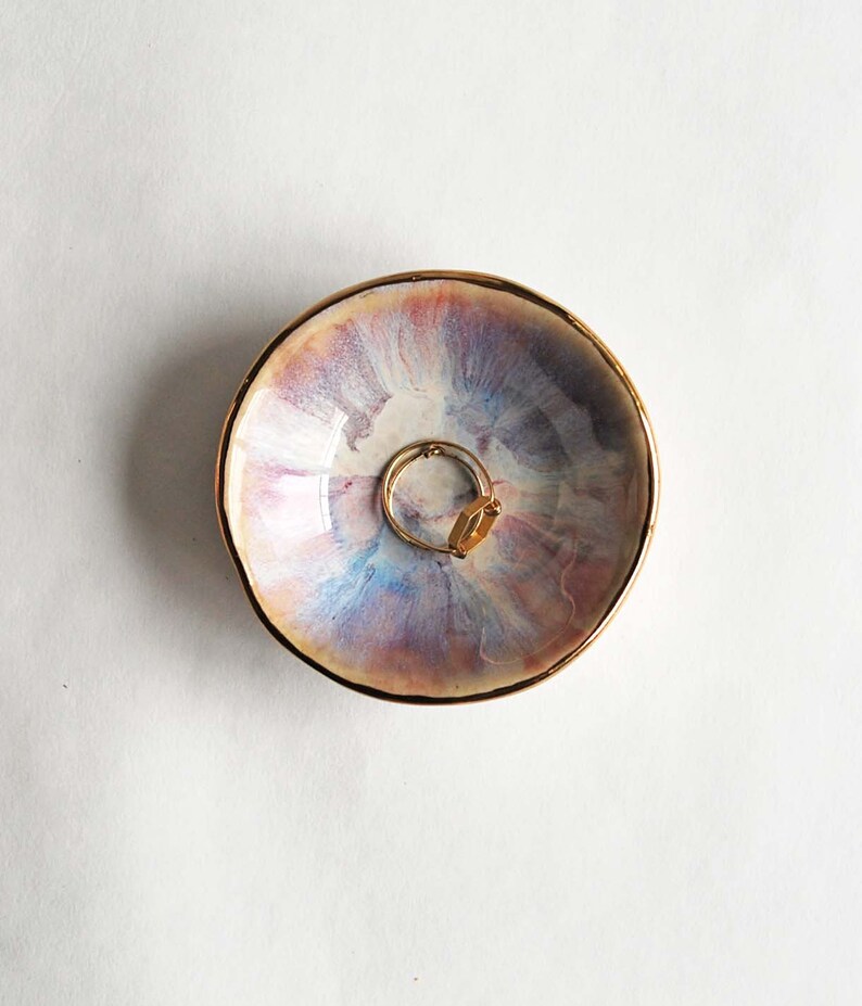 K&ouml;nnte beinhalten: Eine kleine, runde Schmuckschale aus Keramik mit einer schimmernden, irisierenden Glasur in zartem Rosa, Blau und Lavendel. Der Rand der Schale ist mit einem metallisch goldenen Akzent versehen. Ein goldfarbener Ring liegt in der Mitte.